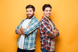 Portrait of a two smiling young men standing with arms folded back to back isolated over yellow background