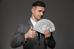 Satisfied lucky chief man 30s in business suit holding fan of cash money dollar currency and showing thumb up isolated over gray background