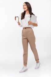 Full length portrait of a happy asian businesswoman carrying laptop computer and cup of coffee to go while walking isolated over white background