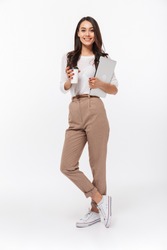 Full length portrait of a smiling asian businesswoman carrying laptop computer and cup of coffee to go while standing isolated over white background