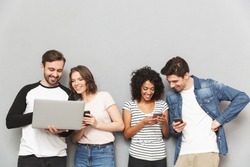 Photo of emotional group of friends standing isolated over grey wall background chatting by mobile phones and laptop computer.