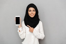 Portrait of young arab woman 20s in islamic headscarf with oriental makeup pointing finger on black screen of cell phone isolated over gray background