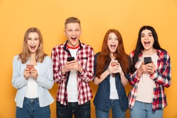 Group of excited school friends holding mobile phones while standing together over yellow background