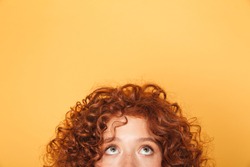 Half face of young curly redhead woman looking up at copy space isolated over yellow background