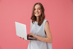Image of cheerful young woman standing isolated over pink background using laptop computer. Looking camera.