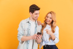 Photo of happy people man and woman laughing while pointing finger at smartphone isolated over yellow background