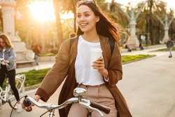 Photo of young pretty woman outdoors walking on bicycle on the street in park holding coffee in hand.