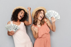Portrait of two happy delighted girls with different type of skin wearing straw hats rejoicing win and dancing while holding fans of cash money isolated over gray background