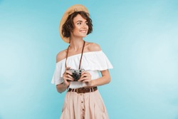 Portrait of a smiling young woman in summer hat standing with photo camera and looking away at copy space isolated over blue background