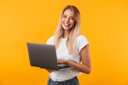 Portrait of a smiling young blonde girl holding laptop computer isolated over yellow background
