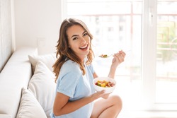Cheerful young woman eating healthy breakfast while sitting on a couch at home