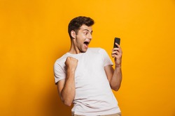 Portrait of an excited young man looking at mobile phone isolated over yellow background, celebrating