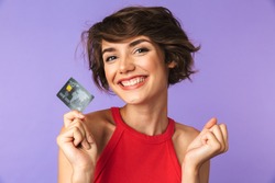 Smiling Pretty brunette woman holding credit card while rejoices and looking at the camera over purple background