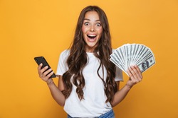 Portrait of an excited young girl with long brunette hair standing over yellow background, holding money banknotes, using mobile phone
