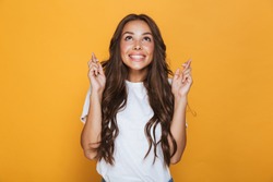 Portrait of a smiling young girl with long brunette hair standing over yellow background, holding fingers crossed for good luck
