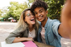 Smiling young multiethnic couple spending time together at the park, studying while sitting at the table and taking a selfie