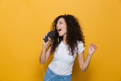 Image of happy woman 20s with curly hair singing while holding smartphone like microphone and listening to music via earphones isolated over yellow background