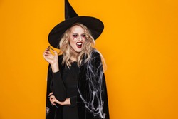 Image of pretty witch woman wearing black costume and halloween makeup smiling at camera isolated over yellow background