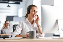 Image of a happy emotional business woman in office callcenter working with computer wearing headphones.