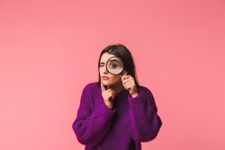 Pretty young girl wearing sweater standing isolated over pink background, looking through a magnifying glass