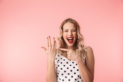 Beautiful excited young blonde woman wearing dress standing isolated over pink background, showing engagement ring on her finger