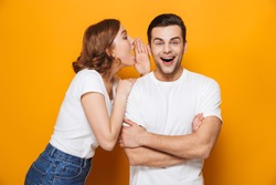 Excited beautiful couple wearing white t-shirts standing isolated over yellow background, telling secret