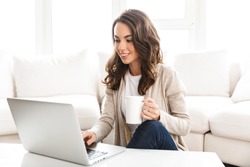 Beautiful young woman working on laptop computer while sitting at the living room, drinking coffee