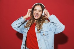 Portrait of a cheerful stylish young woman wearing denim jacket standing isolated over red background, listening to music with headphones, dancing