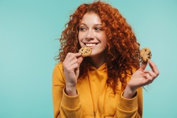 Portrait of alluring redhead woman 20s smiling and eating sweet cookies isolated over blue background