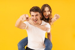Portrait of beautiful couple man and woman in basic t-shirts pointing finger at camera together while doing piggyback ride isolated over yellow background