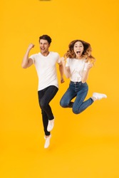 Full length portrait of beautiful couple man and woman in basic t-shirts rejoicing while clenching fists isolated over yellow background