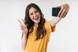 Image of beautiful brunette woman laughing and showing peace sign while taking selfie photo on cellphone isolated over white background