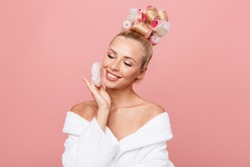 Beautiful smiling young blonde girl wearing bathrobe standing isolated over pink background, using powder puff