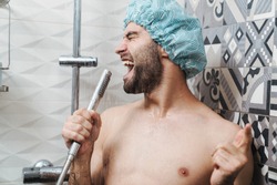 Attractive young cheerful man singing while washing in the shower, wearing shower cap and holding shower head