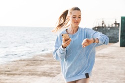 Image of a positive cheerful fitness woman running outdoors on beach using mobile phone listening music with earphones looking at watch clock.