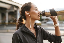 Image of serious athletic woman using earphones and drinking water while working out near road bridge in morning
