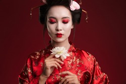 Image of beautiful geisha woman in traditional japanese kimono holding flower isolated over red background