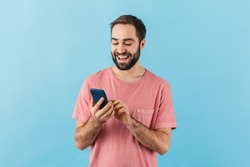 Portrait of a young cheerful excited bearded man wearing t-shirt standing isolated over blue background, using mobile phone