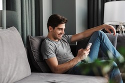 Portrait of an attractive smiling young bearded man wearing casual clothes sitting on a couch at the living room, using mobile phone