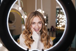 Photo of a young happy cute blonde girl indoors in beauty salon looking at camera through ring light lamp.