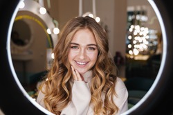 Photo of a young happy cute blonde girl indoors in beauty salon looking at camera through ring light lamp.
