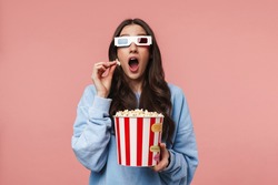 Portrait of a pretty cheerful girl with long curly brunette hair wearing sweatshirt standing isolated over pink background, wearing 3d glasses, holding popcorn bucket
