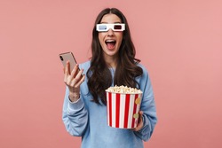 Portrait of a pretty cheerful girl with long curly brunette hair wearing sweatshirt standing isolated over pink background, wearing 3d glasses, holding popcorn bucket, holding mobile phone