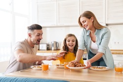 Photo of happy caucasian beautiful family smiling and eating croissants while having breakfast in modern kitchen