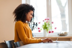 Image of cheerful african american woman using laptop while sitting at table in living room