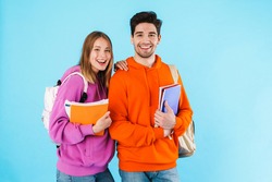 Portrait of a cheerful young couple of students wearing backpacks, carrying textbooks standing isolated over blue background