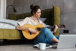 Image of young caucasian brunette woman sitting on floor and playing acoustic guitar at home