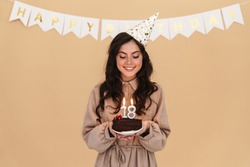 Image of happy young woman in party cone smiling while posing with birthday cake isolated over beige background