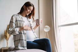 Beautiful happy pregnant woman smiling and drinking coffee while sitting on couch at home