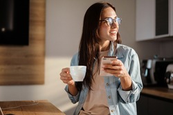 Happy charming woman using smartphone and drinking coffee at home kitchen
